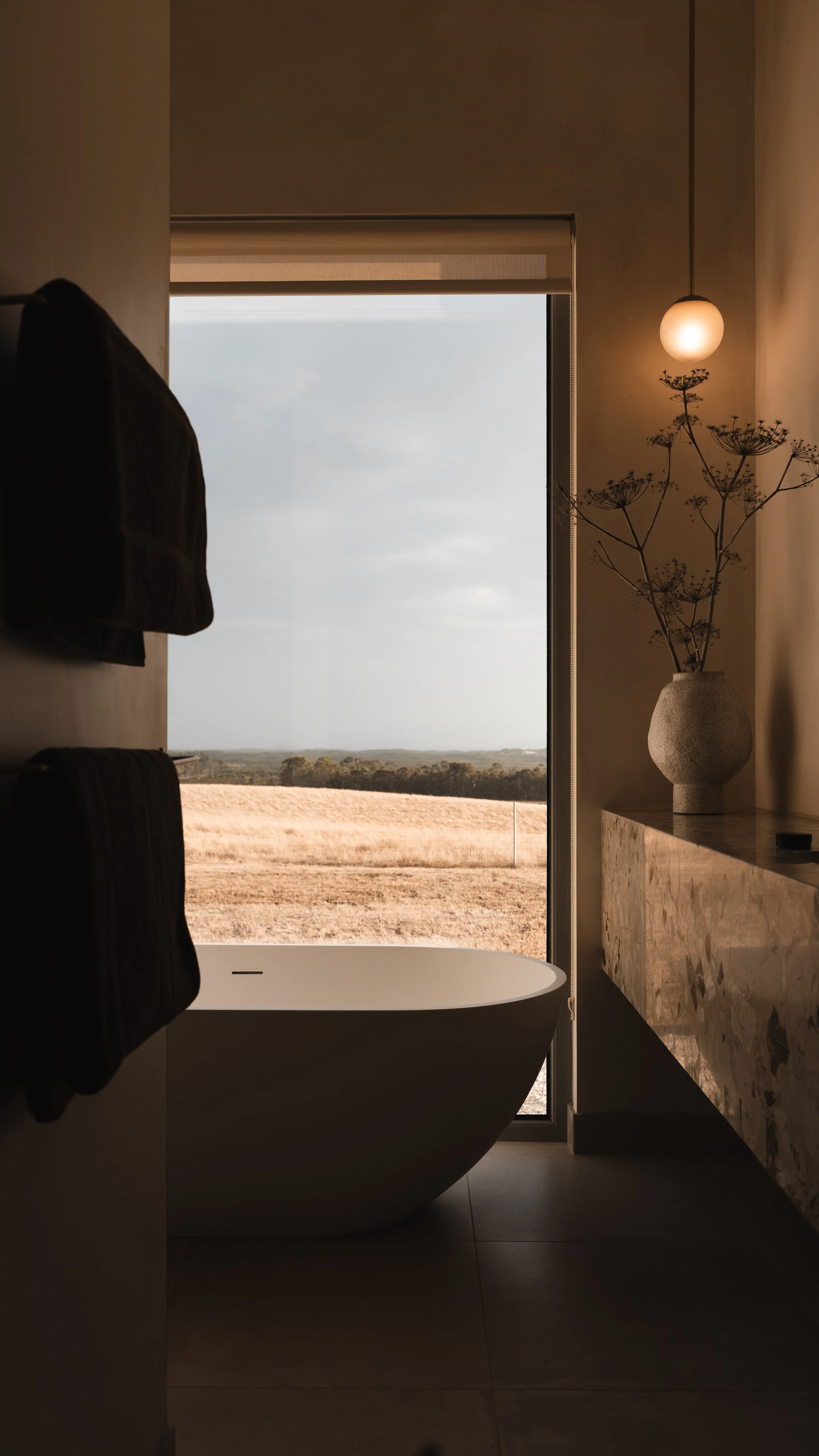 Modern bathroom at Box Hill Esperance with a freestanding bathtub near a window overlooking a field, with a vase and dried flowers on a marble countertop, and warm lighting from a pendant lamp.