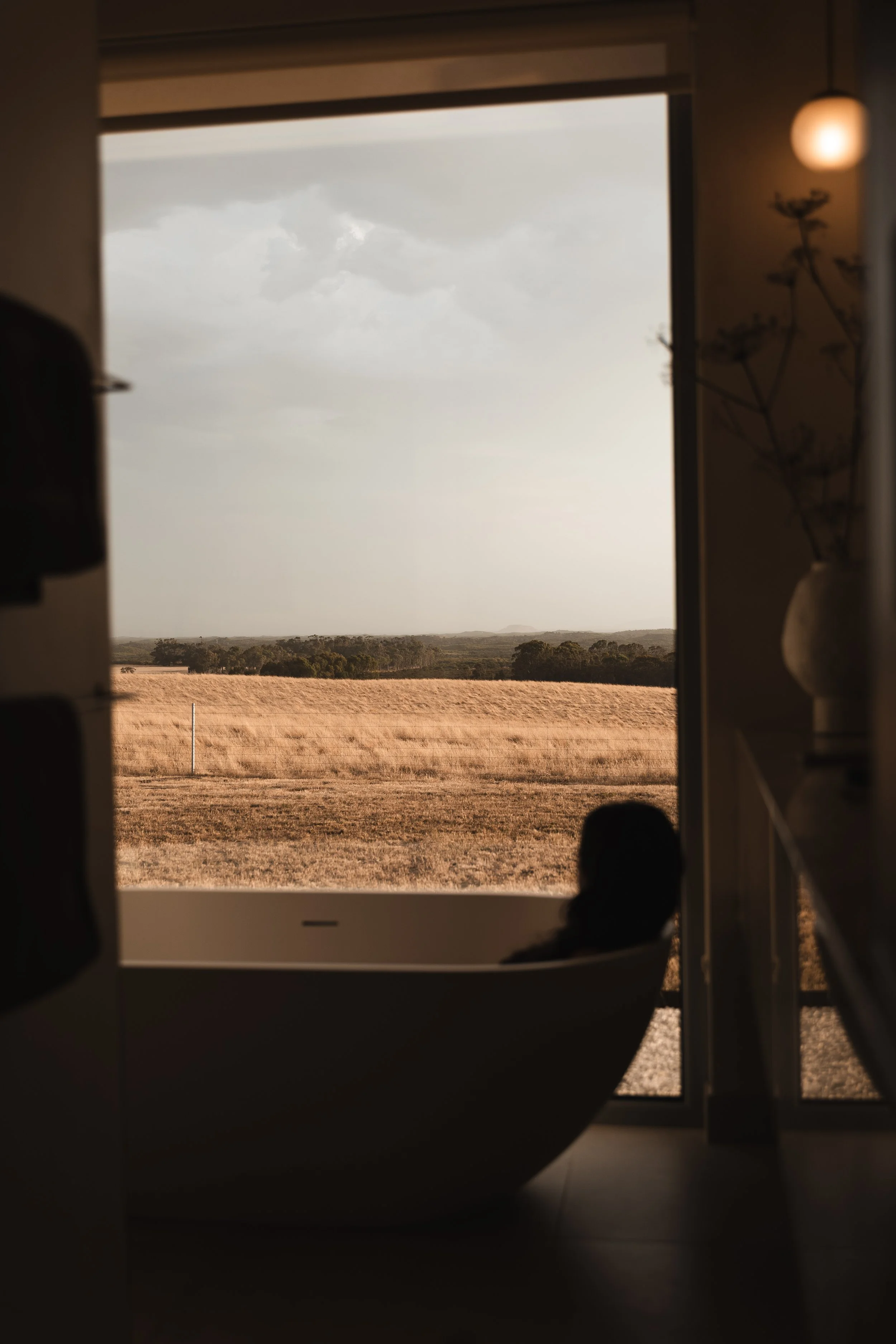 Person relaxing in a bathtub looking out at an open field and cloudy sky through a large window.