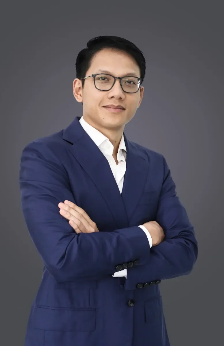 A man wearing glasses and a navy blue suit with a white shirt, standing with arms crossed against a gray background.