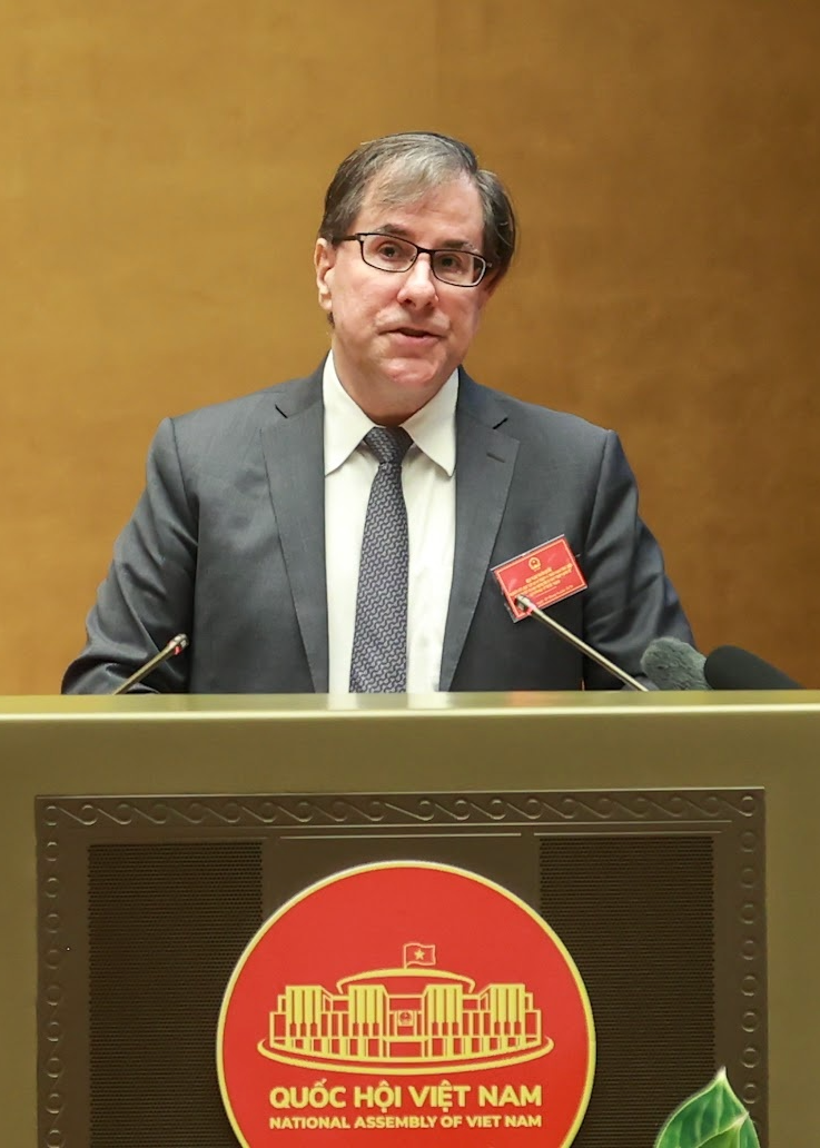 A man in a gray suit and glasses standing at a podium with the emblem of the National Assembly of Vietnam, speaking into microphones.