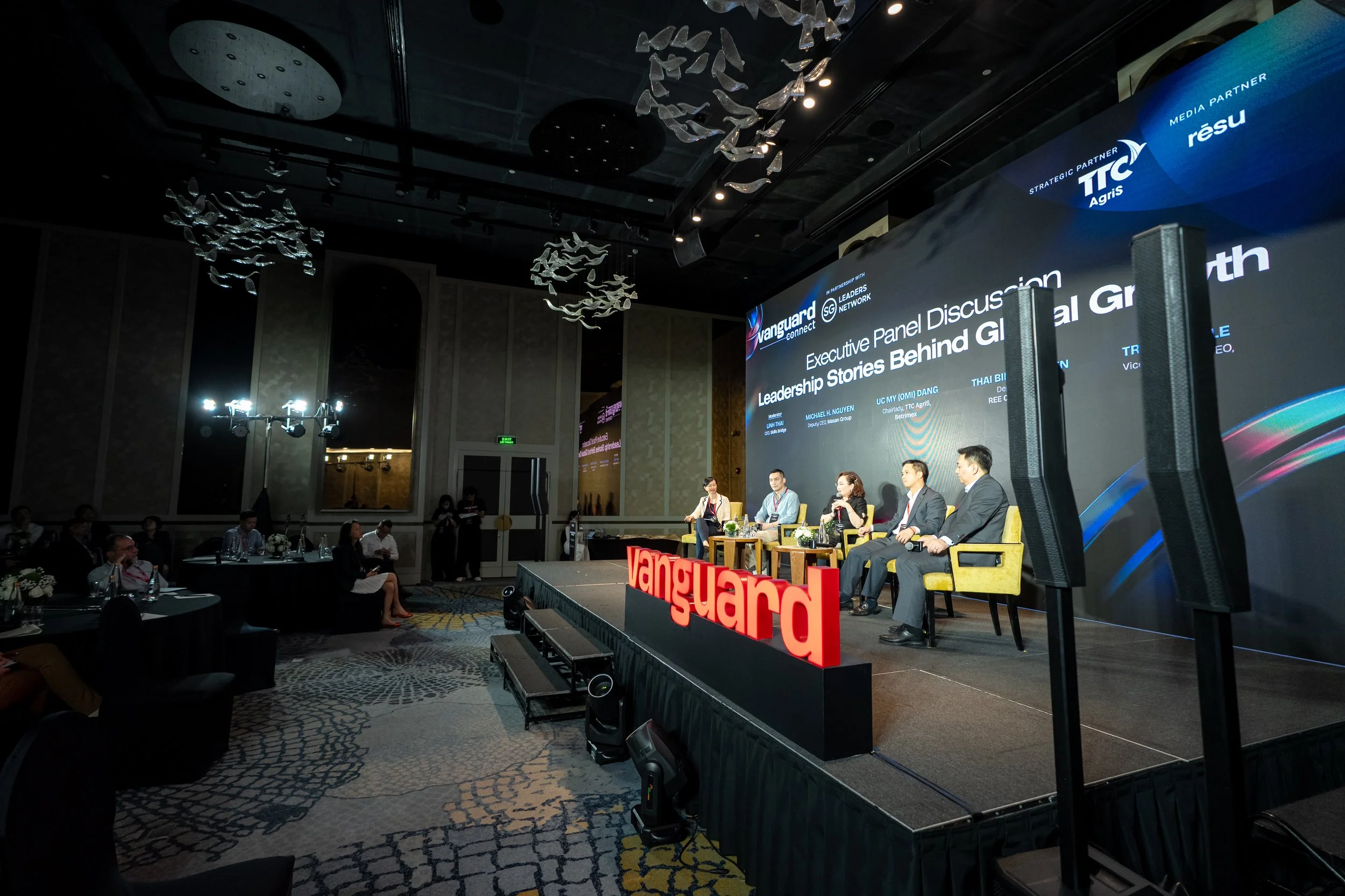 Panel discussion at a conference with five speakers seated on stage, a large screen behind them displaying the event details, and an audience in the foreground. The stage features a red 'Vanguard' sign.