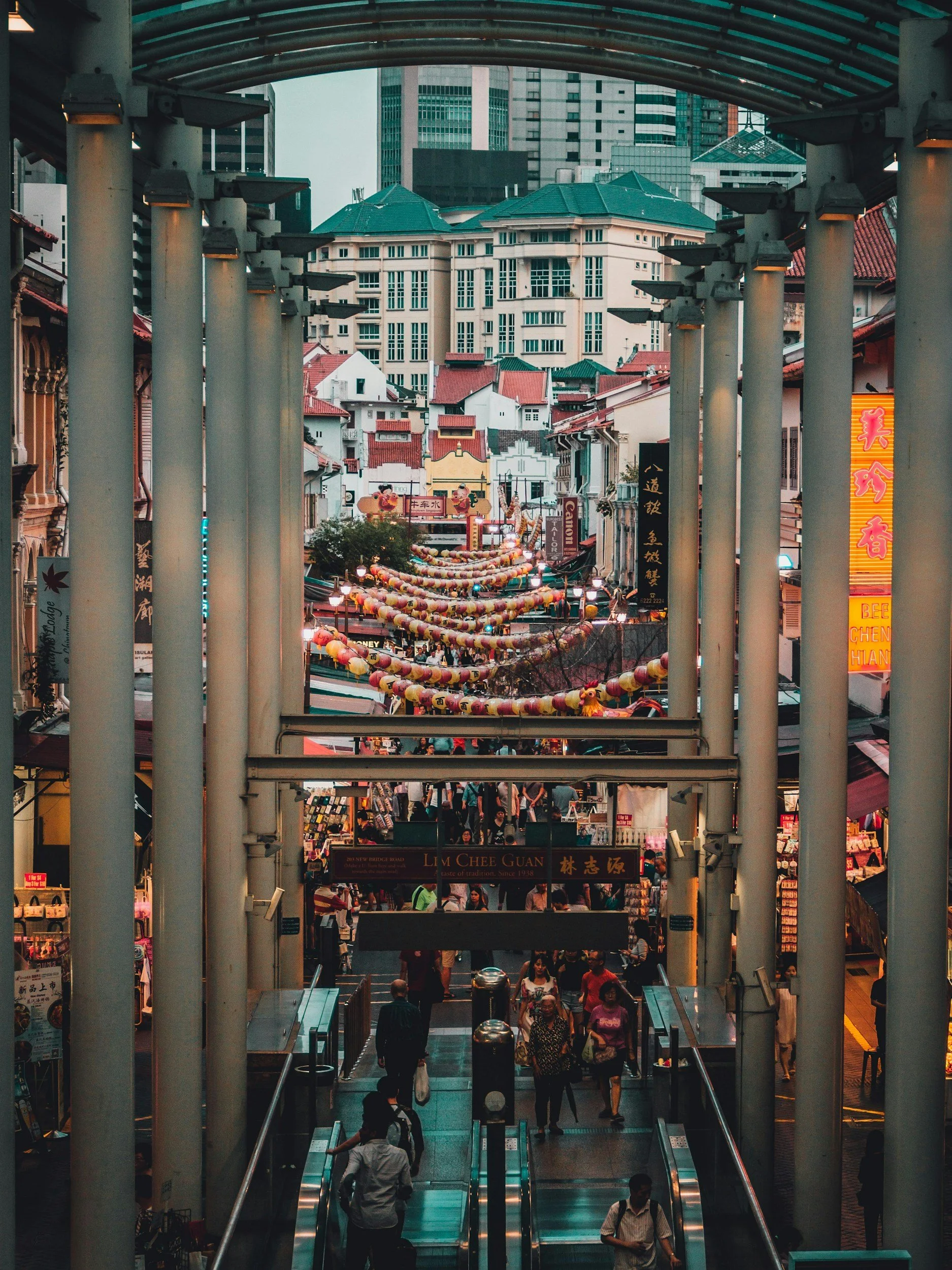 View of a busy street market in Chinatown, Singapore, seen from an elevated walkway with columns, with colorful lanterns hanging across the street and tall modern buildings in the background.
