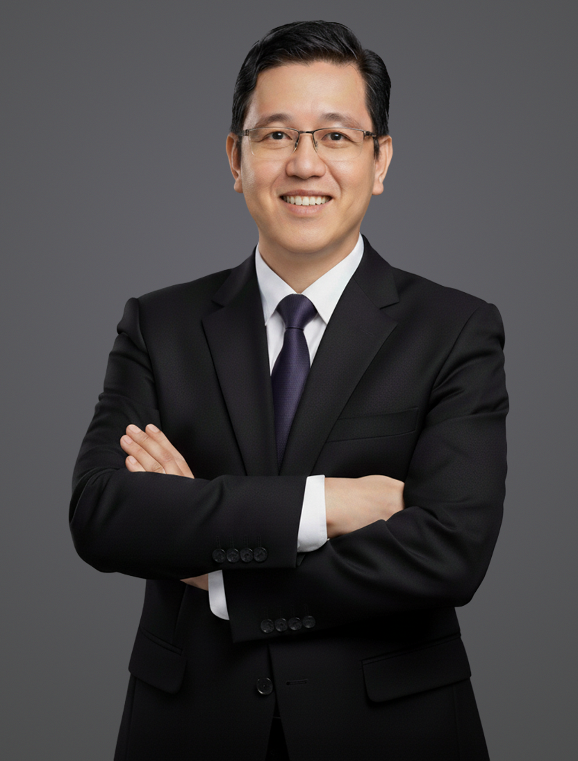 A smiling man in a black suit, white shirt, and dark tie, standing with arms crossed against a gray background.