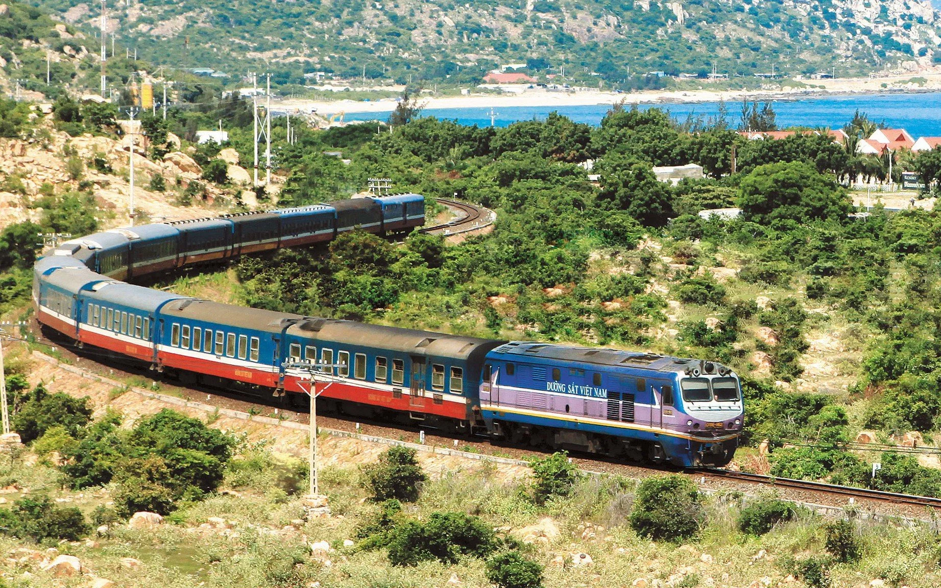 A colorful train travels through lush green hills near the coast, with blue water and distant houses in the background.