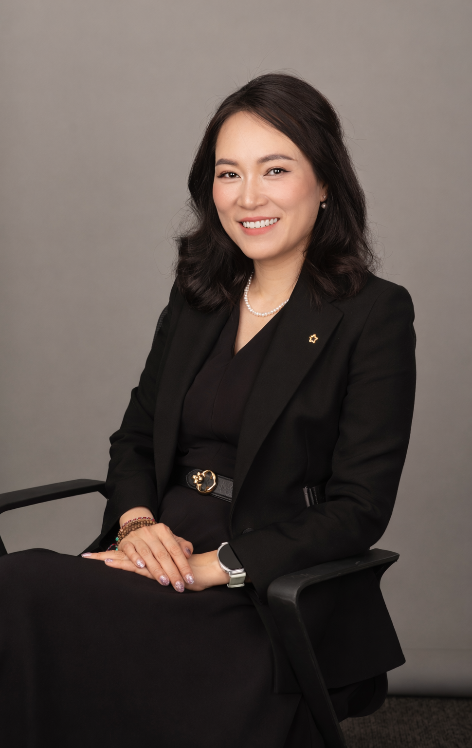 Professional woman in black blazer smiling, sitting in a black chair against a gray background.