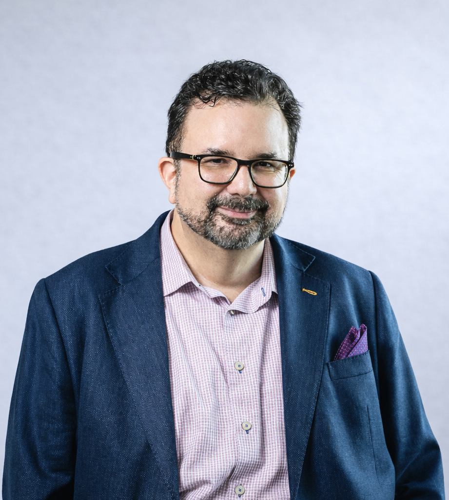 Portrait of a man with dark hair and glasses, wearing a navy blazer and a pink checkered shirt, standing against a light grey background.