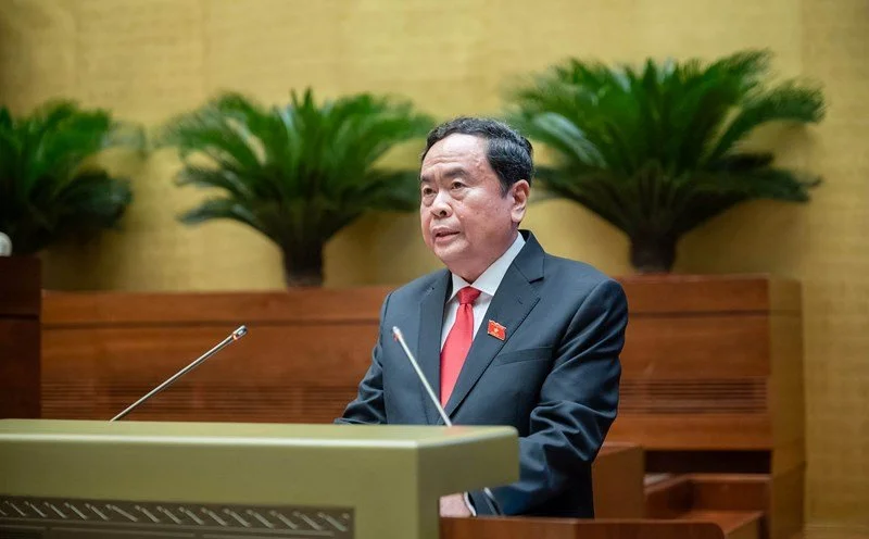 A man in a black suit with a red tie and lapel pin standing at a podium, speaking, in front of a wooden wall with potted plants in the background.