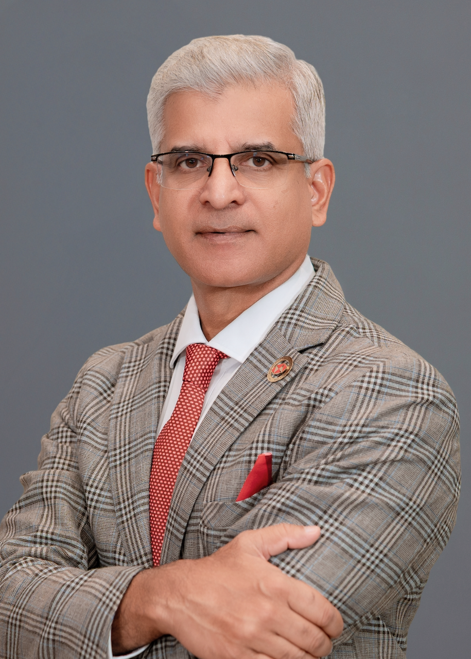 A middle-aged man with gray hair, wearing glasses, a plaid blazer, white shirt, red dotted tie, and a red pocket square, standing against a plain gray background.