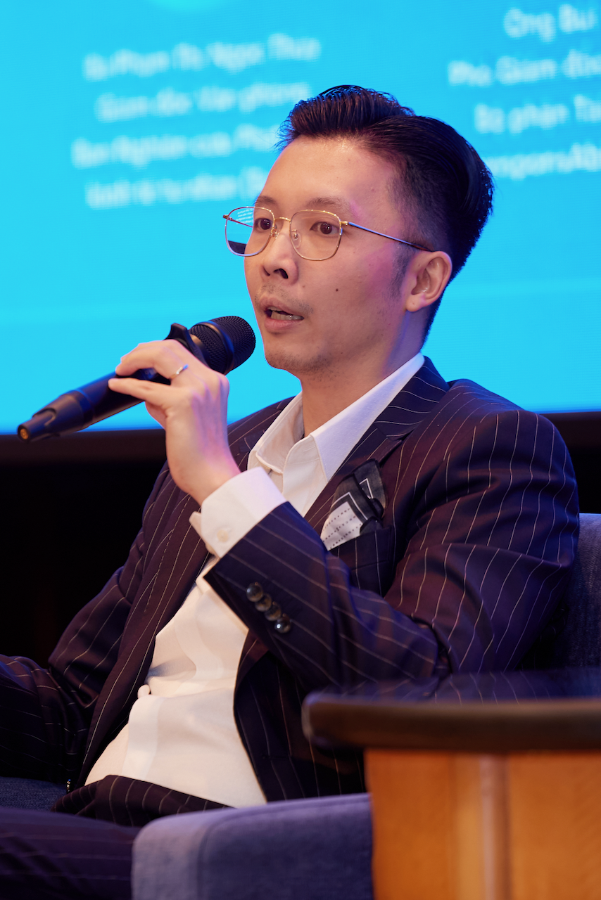 Man in pinstripe suit and glasses speaking into a handheld microphone during a panel or presentation.