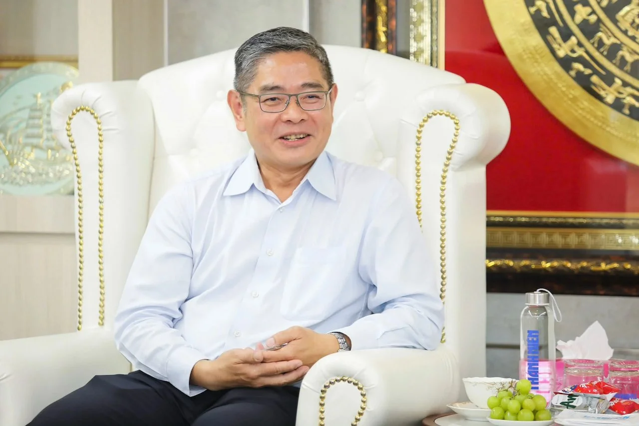 A man with glasses wearing a white shirt, sitting on a white ornate chair with gold detail, smiling. There are grapes, a water bottle, and snacks on a table nearby.