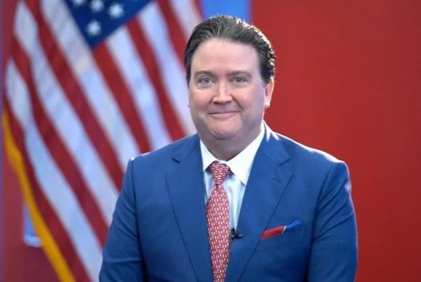 A man in a blue suit with a pink patterned tie and a white shirt, standing in front of American flags, smiling with a red background.