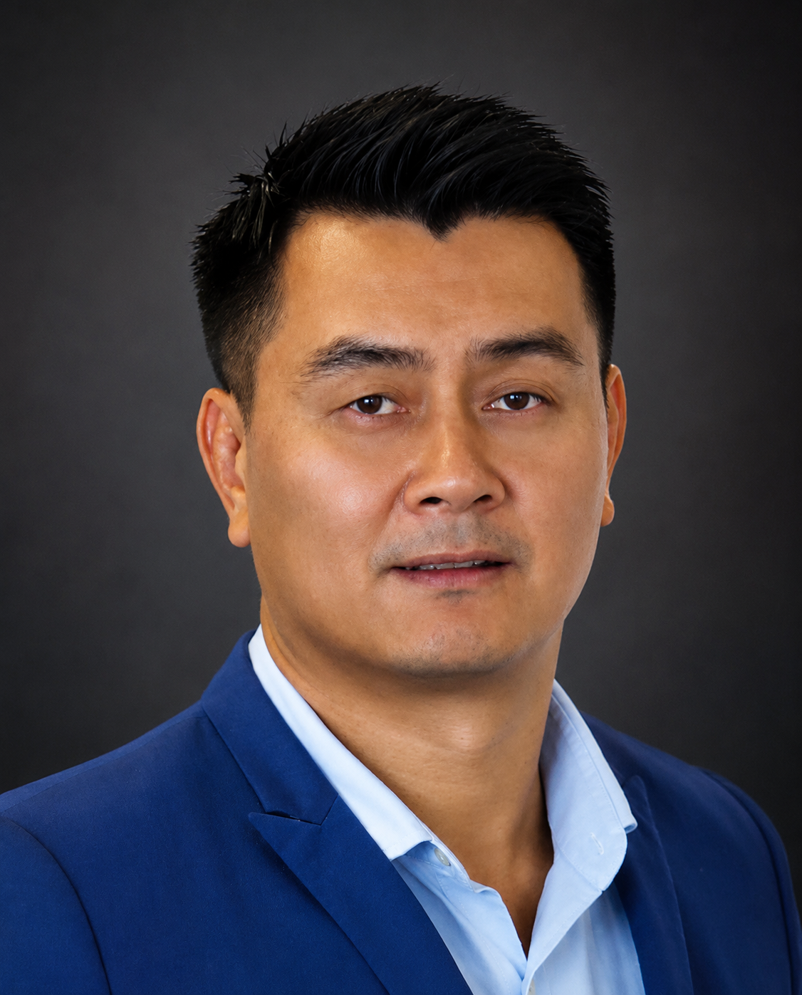 A professional headshot of a man with short black hair, wearing a blue suit jacket and a light blue dress shirt, against a dark background.