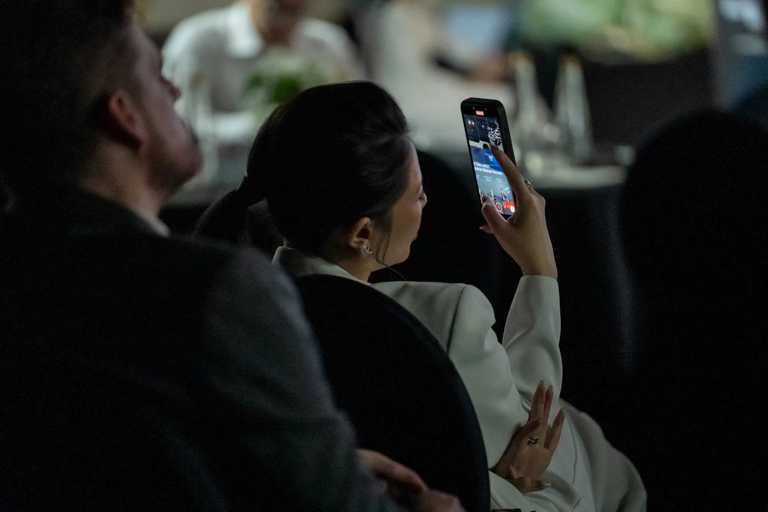 A woman in a white blazer sitting in a dark room, holding a smartphone and taking a photo of a presentation on a screen. A man is seated next to her, looking ahead.