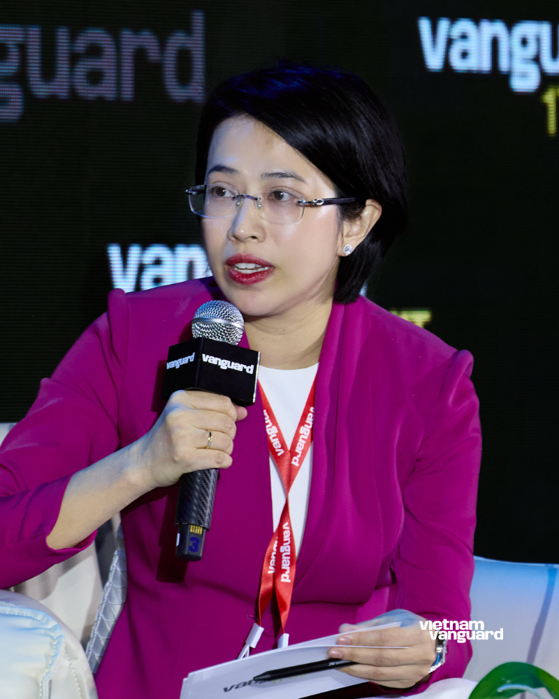 A woman with short black hair, glasses, and wearing a magenta blazer, speaking into a microphone at a Vietnam Vanguard event.