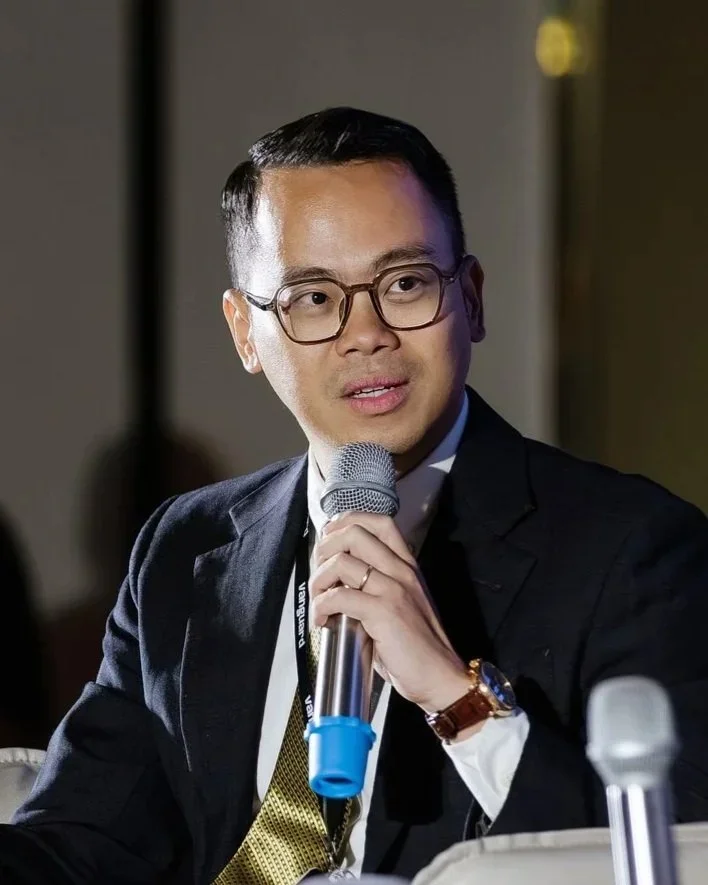 Man in glasses speaking, wearing a blue lanyard and a gray blazer, indoors with blurred background of people and ceiling lights.