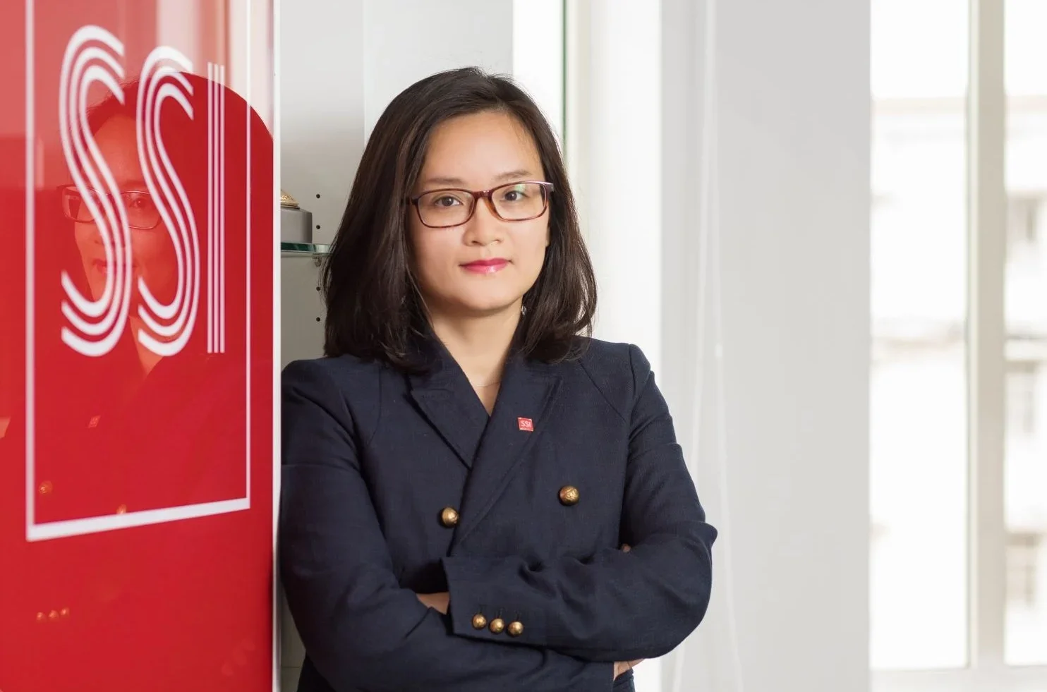 A woman with shoulder-length dark hair and glasses standing with arms crossed, wearing a dark navy blazer with gold buttons, standing near a red door or wall with the letters 'SS' on it in an office setting with large windows in the background.