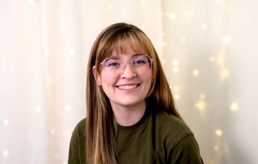 Smiling person with glasses and long brown hair, wearing an olive green top, against a backdrop of twinkling lights.