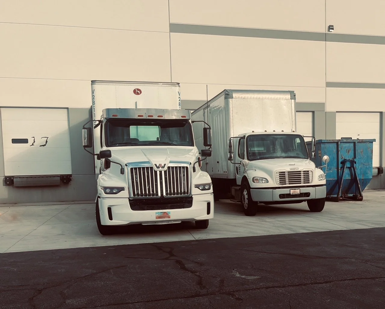 Two white delivery trucks parked in front of a warehouse wall with loading docks. One truck has a larger cab and grill, while the smaller truck is a box truck. There are blue dumpsters on the right side of the image.