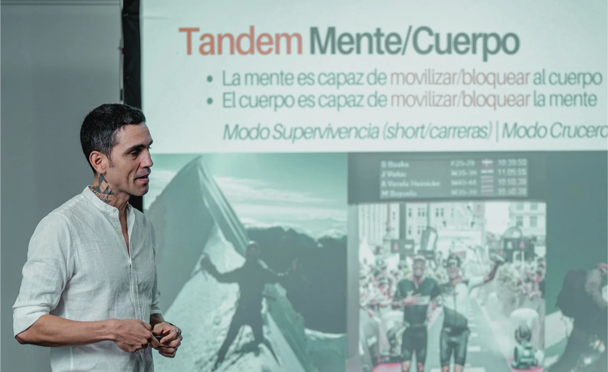 Hombre en una conferencia frente a una diapositiva que dice "Tandem Mente/Cuerpo" y explica que la mente puede movilizar o bloquear el cuerpo y viceversa, en modo supervivencia o modo cruzar.