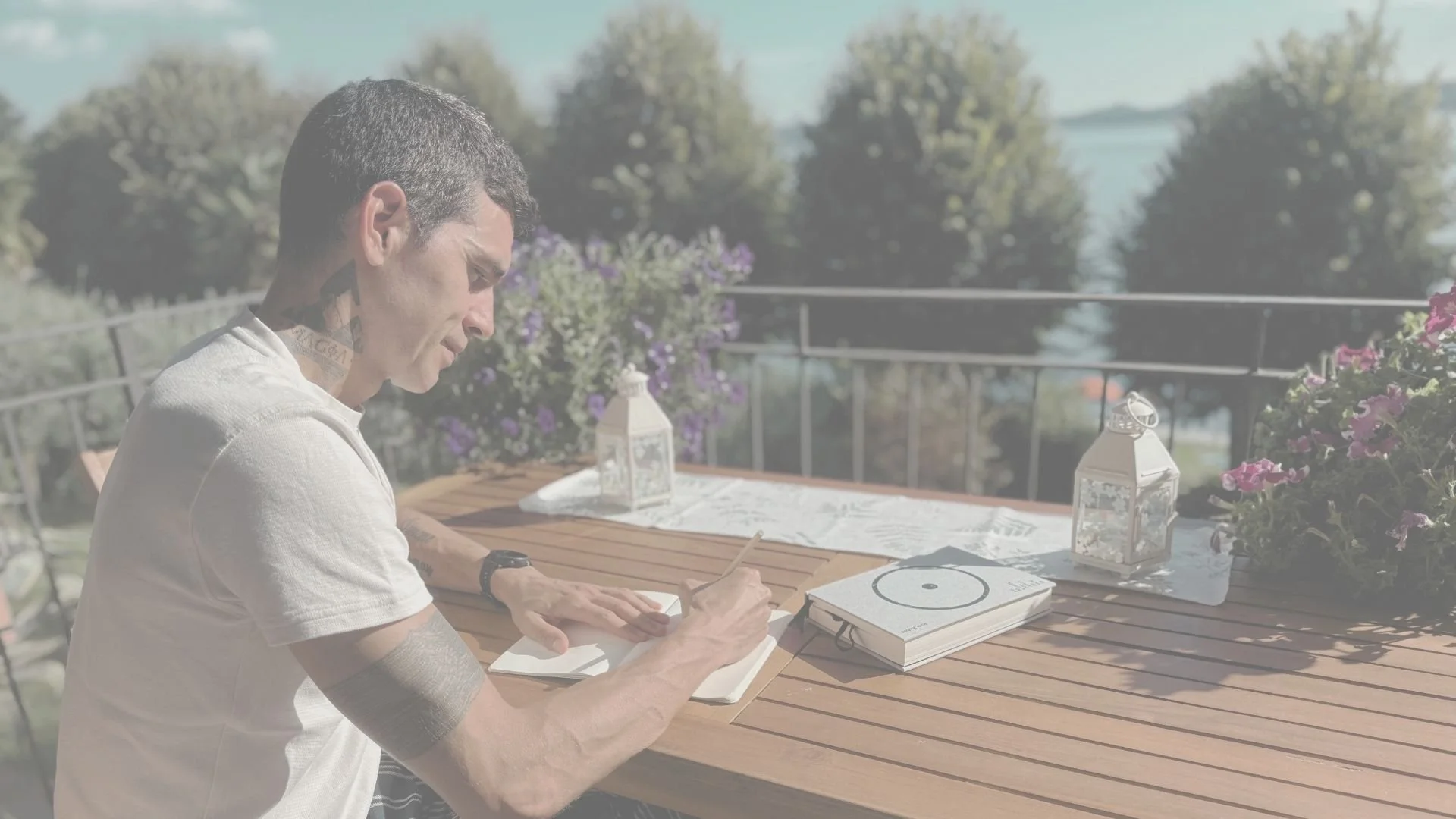 Joven con tatuajes escribiendo en un cuaderno en una mesa de madera en un balcón con flores y vista al mar
