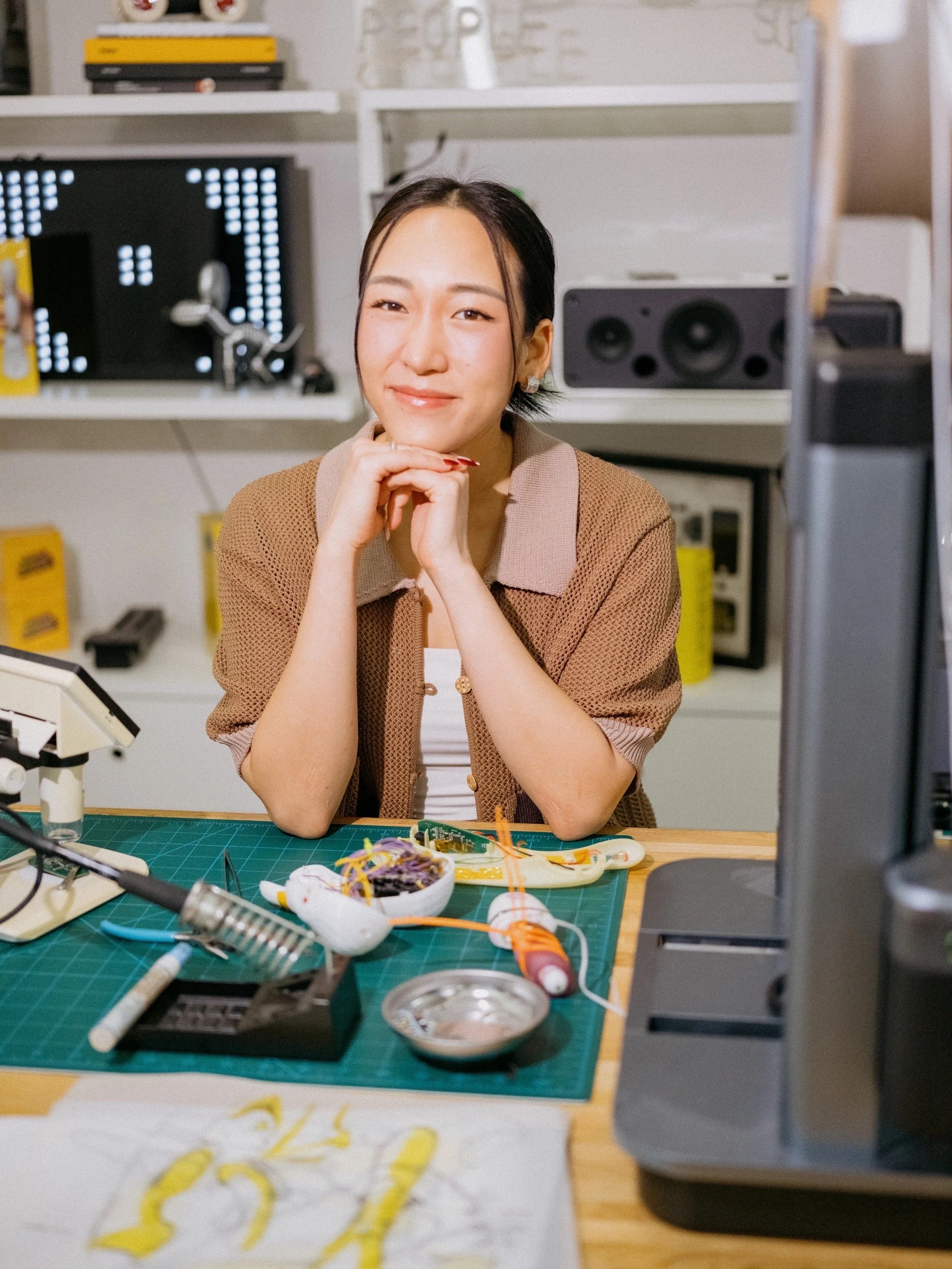 Woman sitting at a cluttered desk with various electronic parts and tools, smiling and resting her chin on her hands in a workspace.