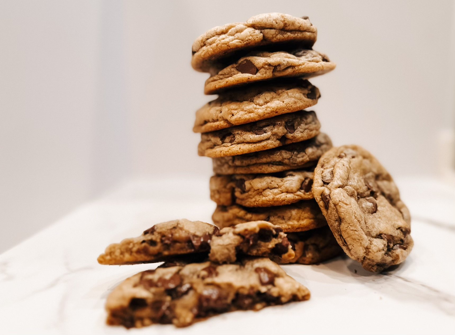 A tall stack of gooey chocolate chip cookies with golden edges and soft centers, artfully arranged on a white surface. A few broken cookie
