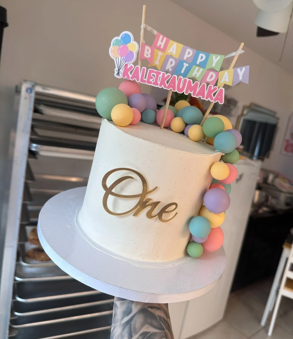 Pastel balloon-themed first birthday cake with smooth buttercream finish, gold ‘One’ topper, and rainbow balloon garland details — handcrafted by Twice As Sweet OC in Huntington Beach