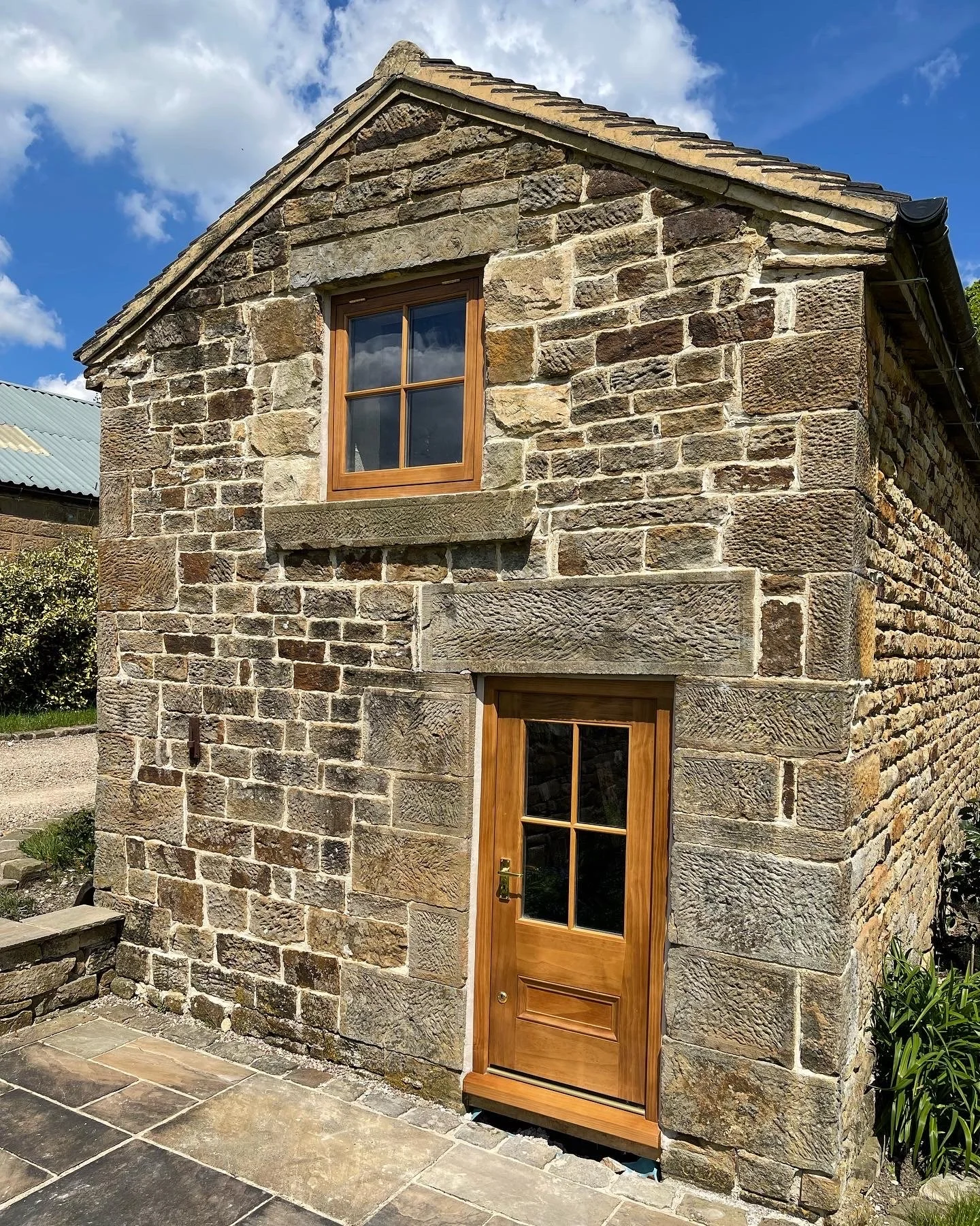 A small stone house with a wooden Accoya timber door and window, situated outside on a sunny day with a blue sky and clouds.