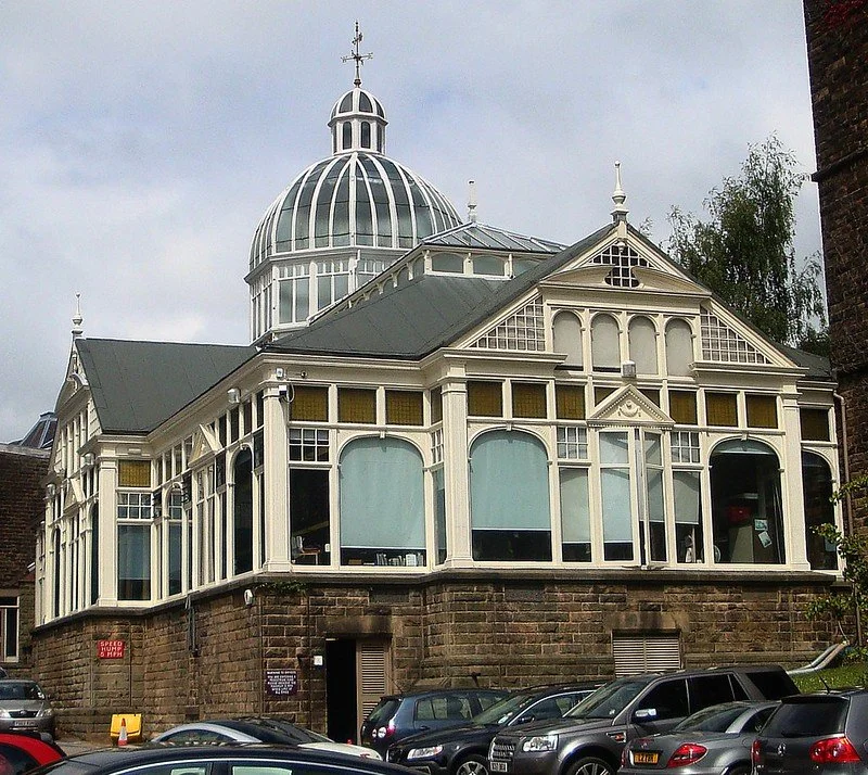 A historic building with ornate white windows and a large glass dome on top, situated on a stone base with parked cars in the foreground.
