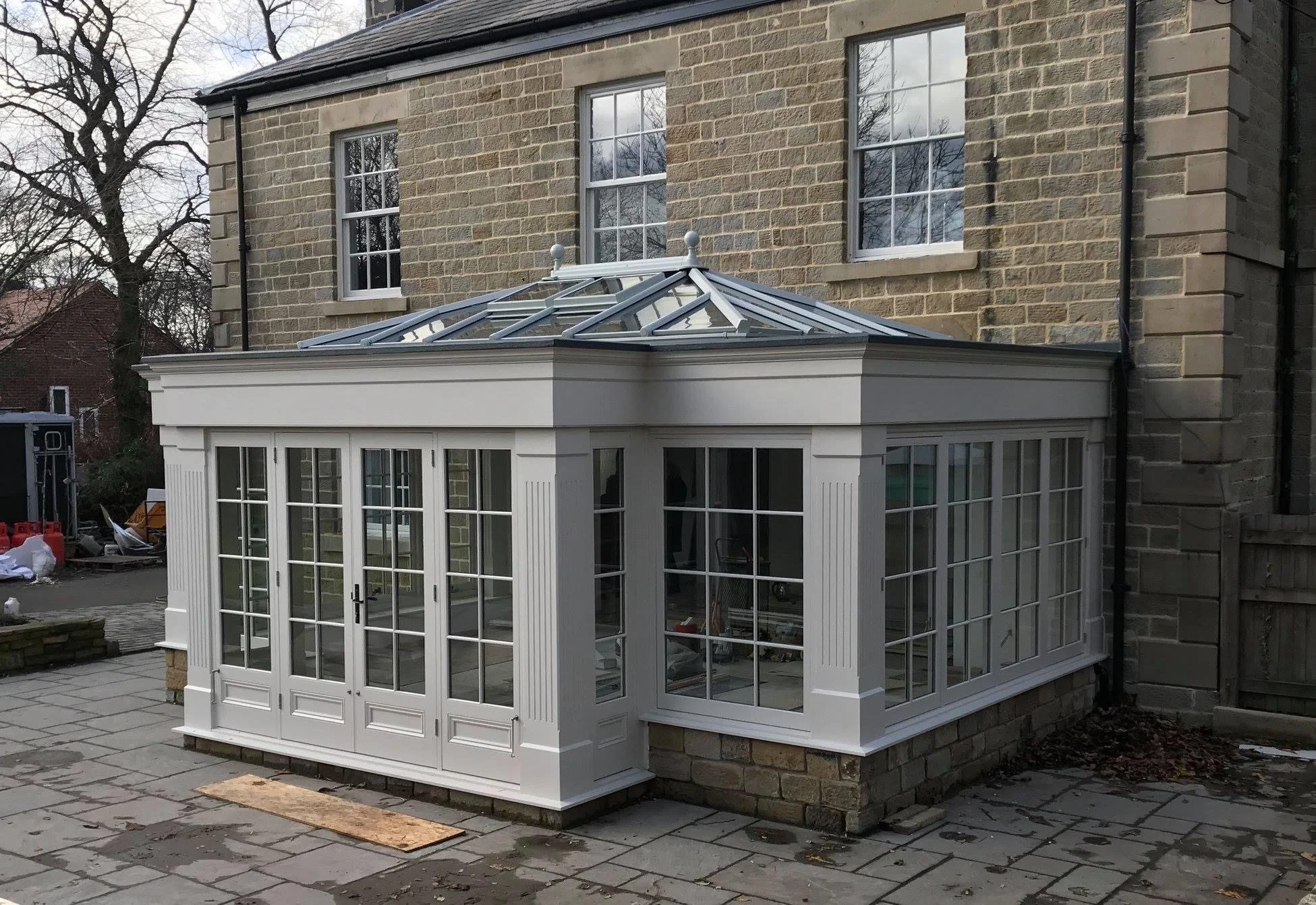 A white conservatory with glass windows and doors attached to a brick house, with a glass roof and decorative finials on top.