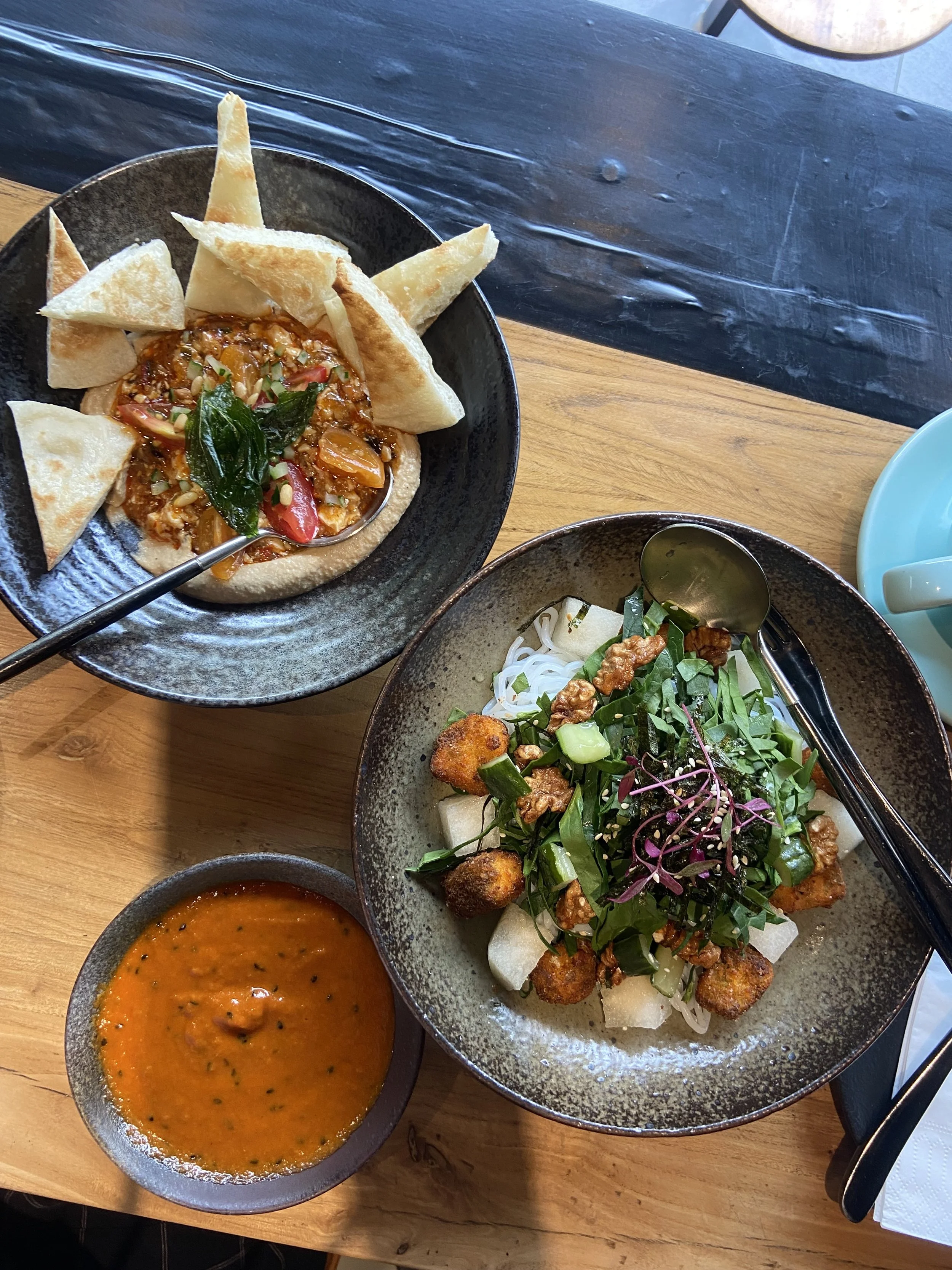 Three Asian dishes on a wooden table: a bowl of curry with naan and papadum, a bowl of soup, and a bowl of noodle salad with greens and fried tofu.