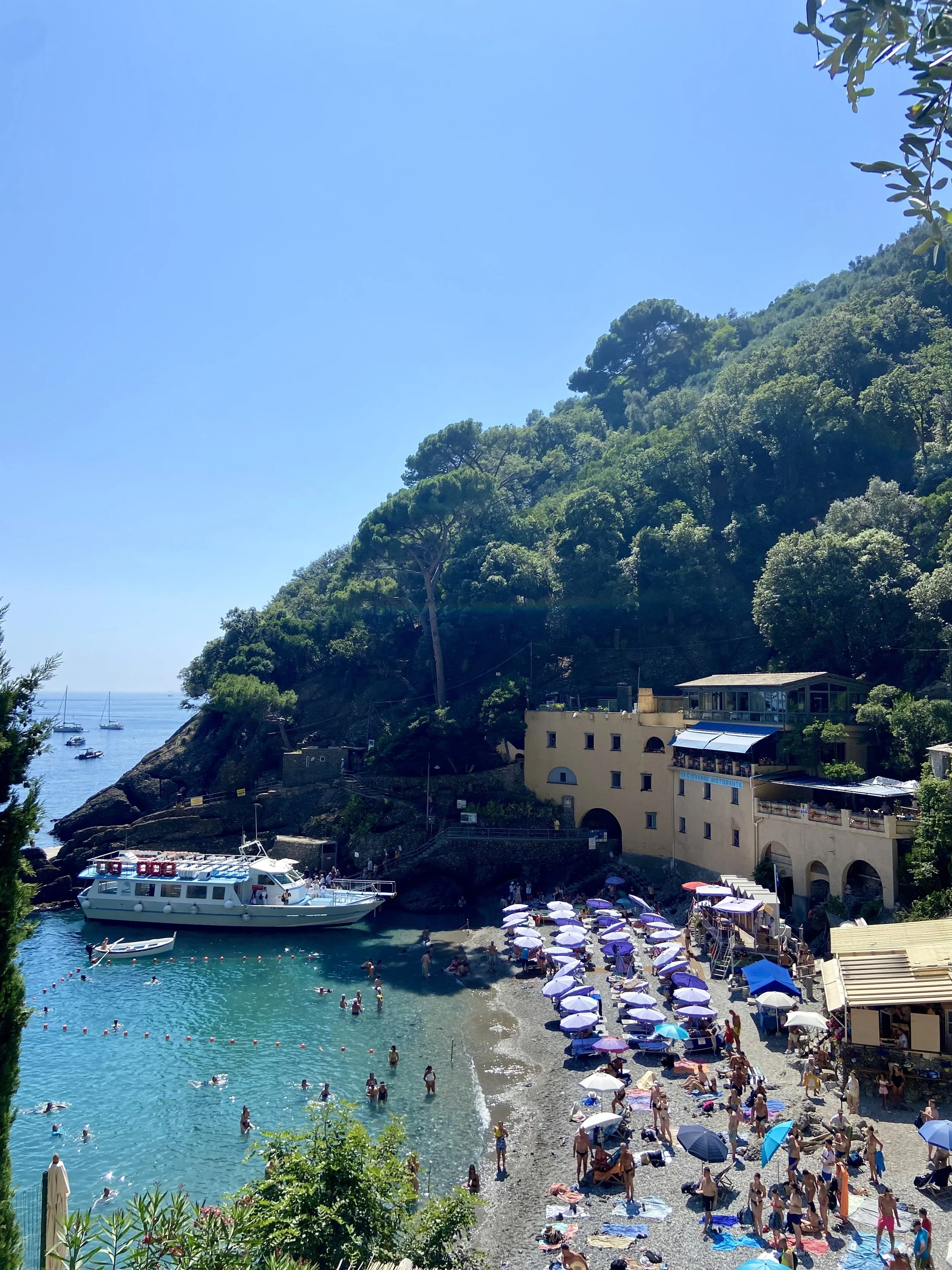 A crowded beach with people sunbathing under umbrellas, swimming and wading in the water, with boats anchored offshore, and a building set into a hillside surrounded by green trees.