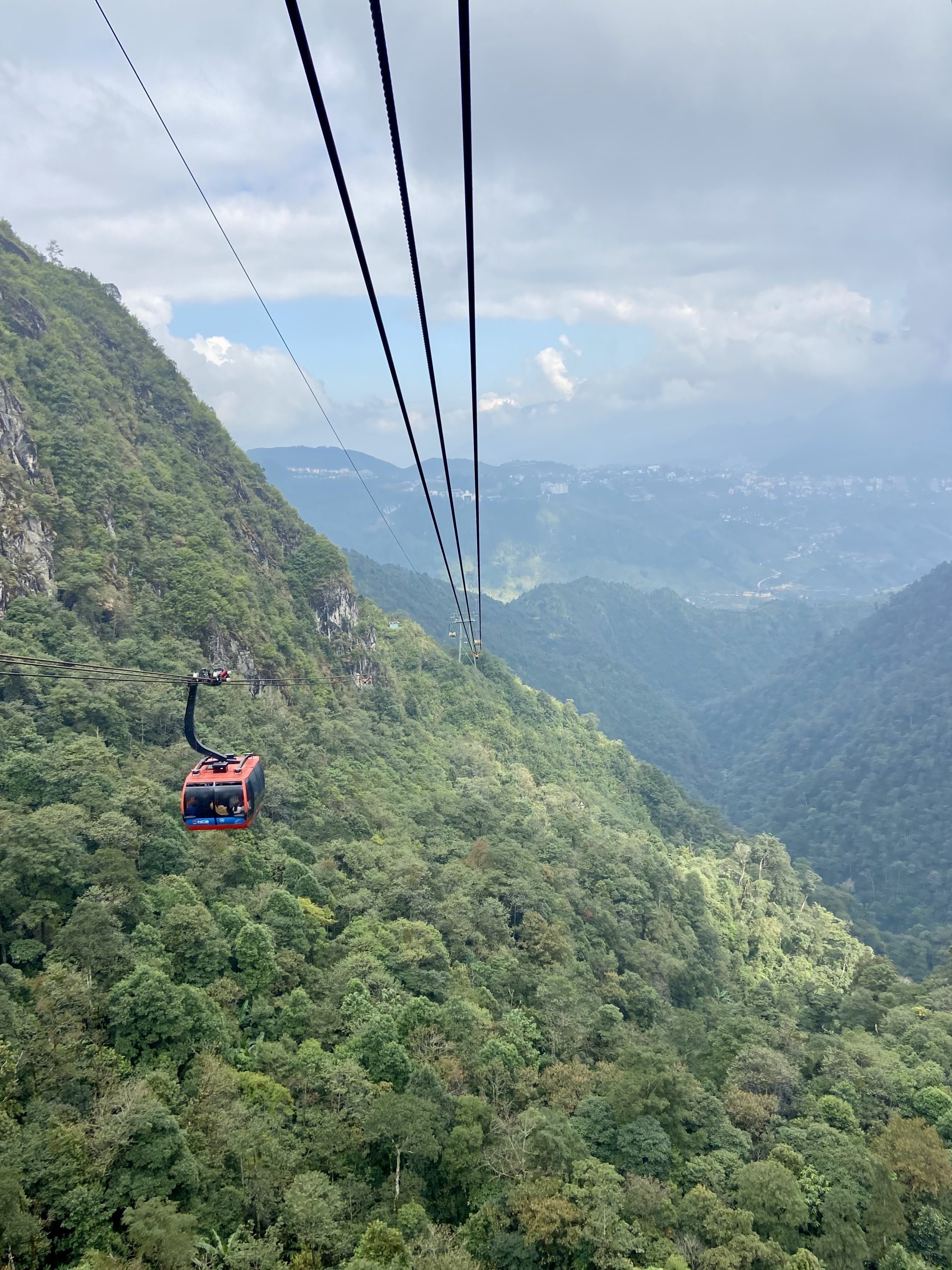 A red cable car is traveling over a lush green mountainous landscape with hills and a cloudy sky in the background.