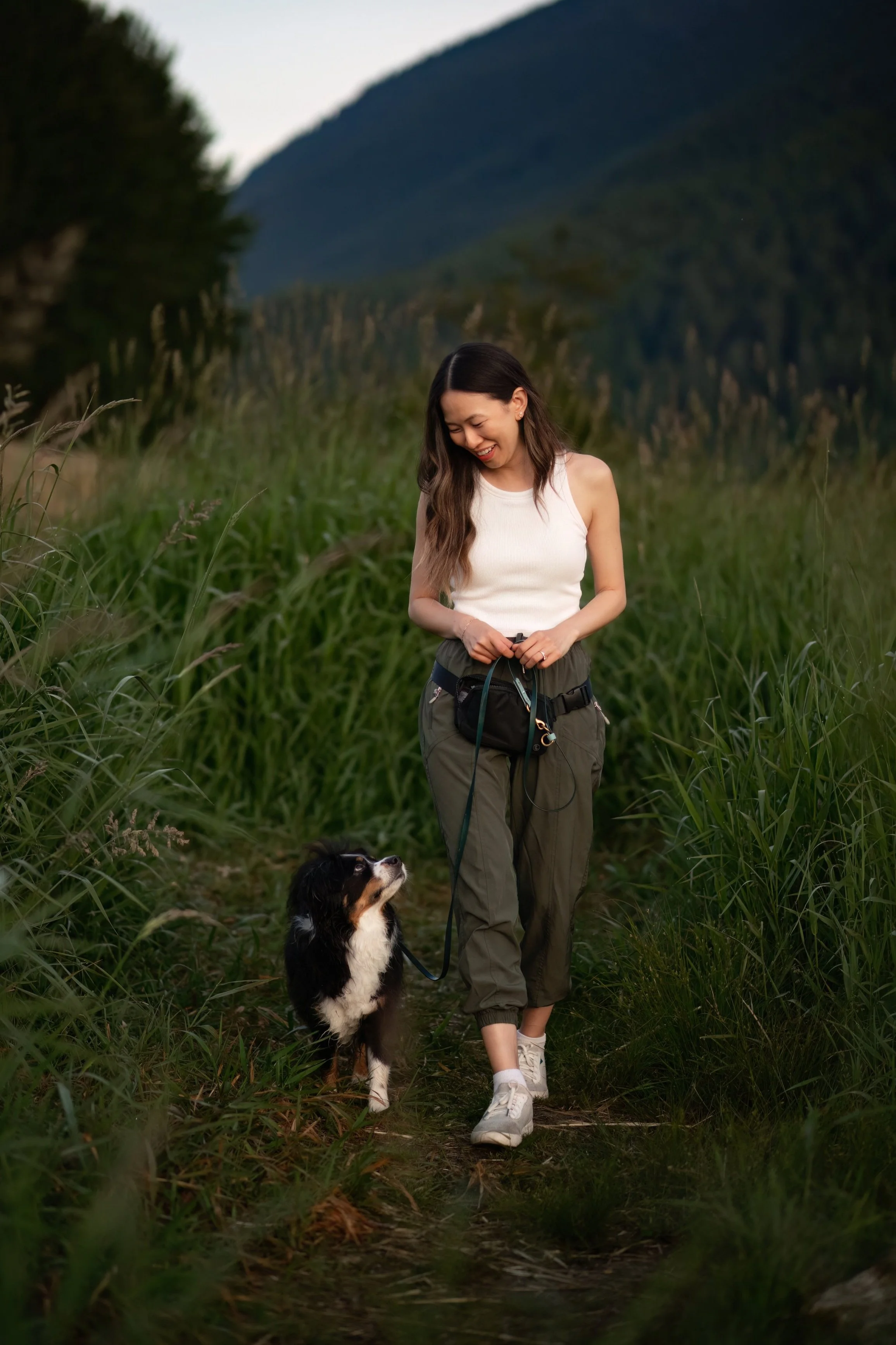 A woman walking a black and white dog on a narrow trail through tall green grass with mountains in the background, smiling and looking down at the dog.
