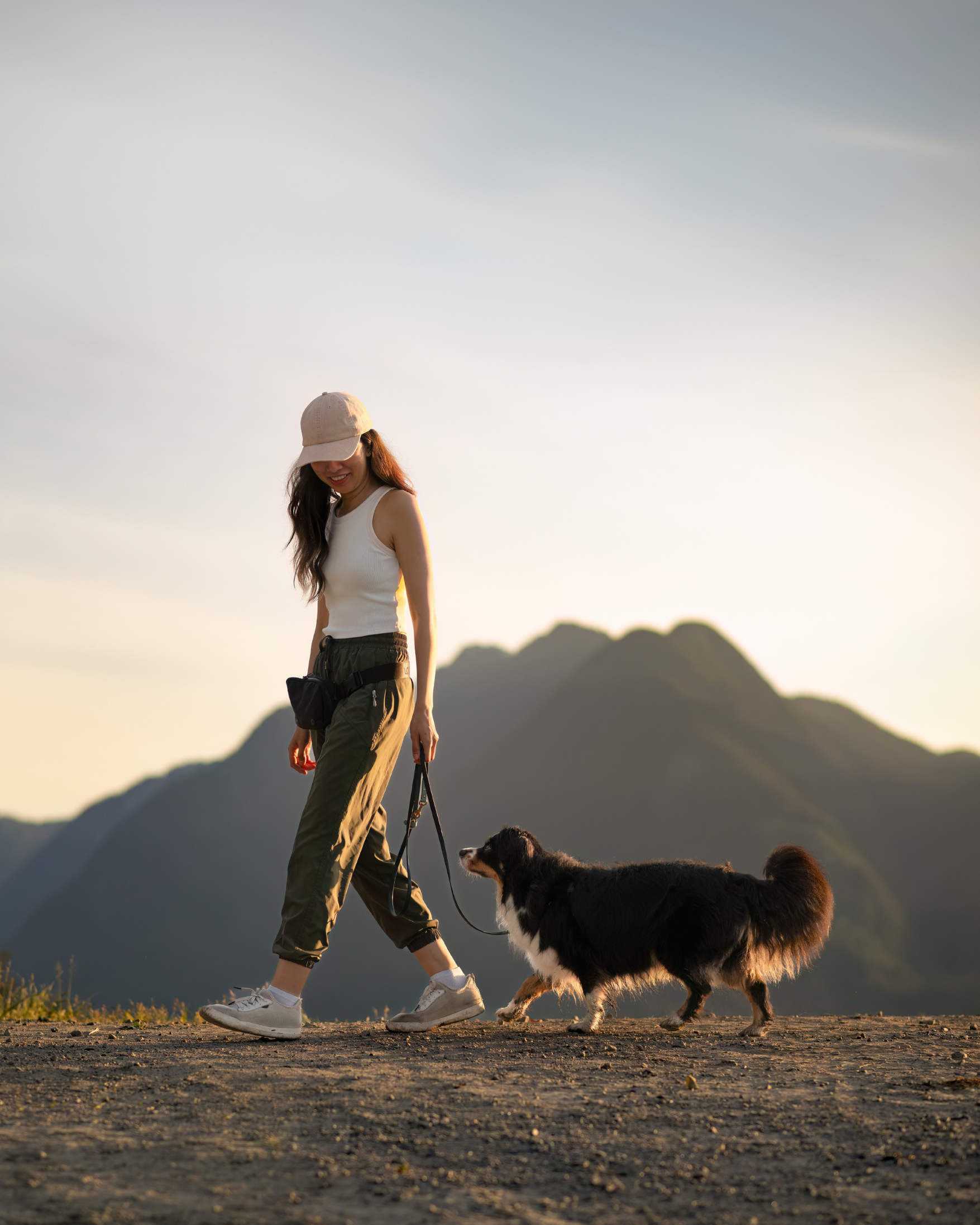 A woman walking her dog outdoors during sunset with mountains in the background.
