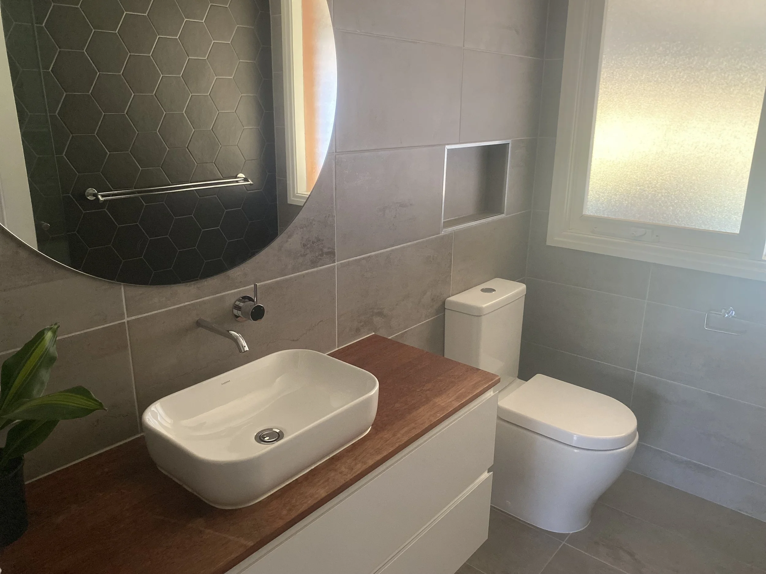 Modern bathroom with gray tiled walls, a white toilet, a wooden countertop with a white vessel sink, and a round mirror. There is a frosted window and a built-in shelf in the wall.