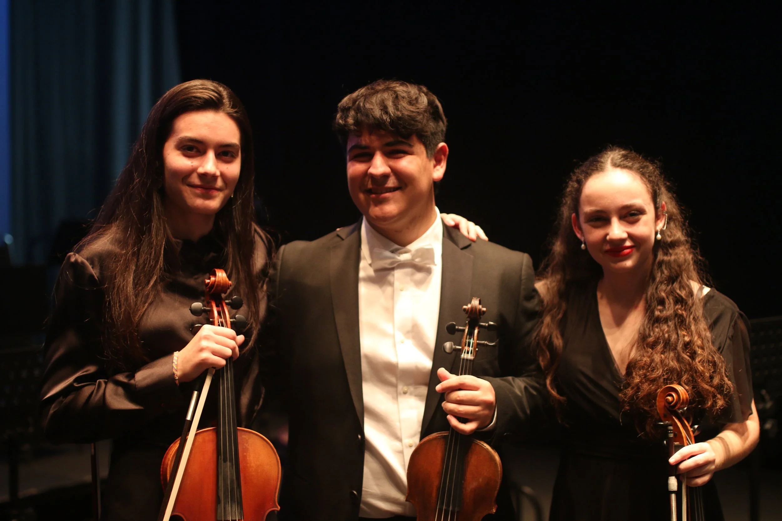 Tres músicos con violines y un director en un escenario oscuro post-concierto.