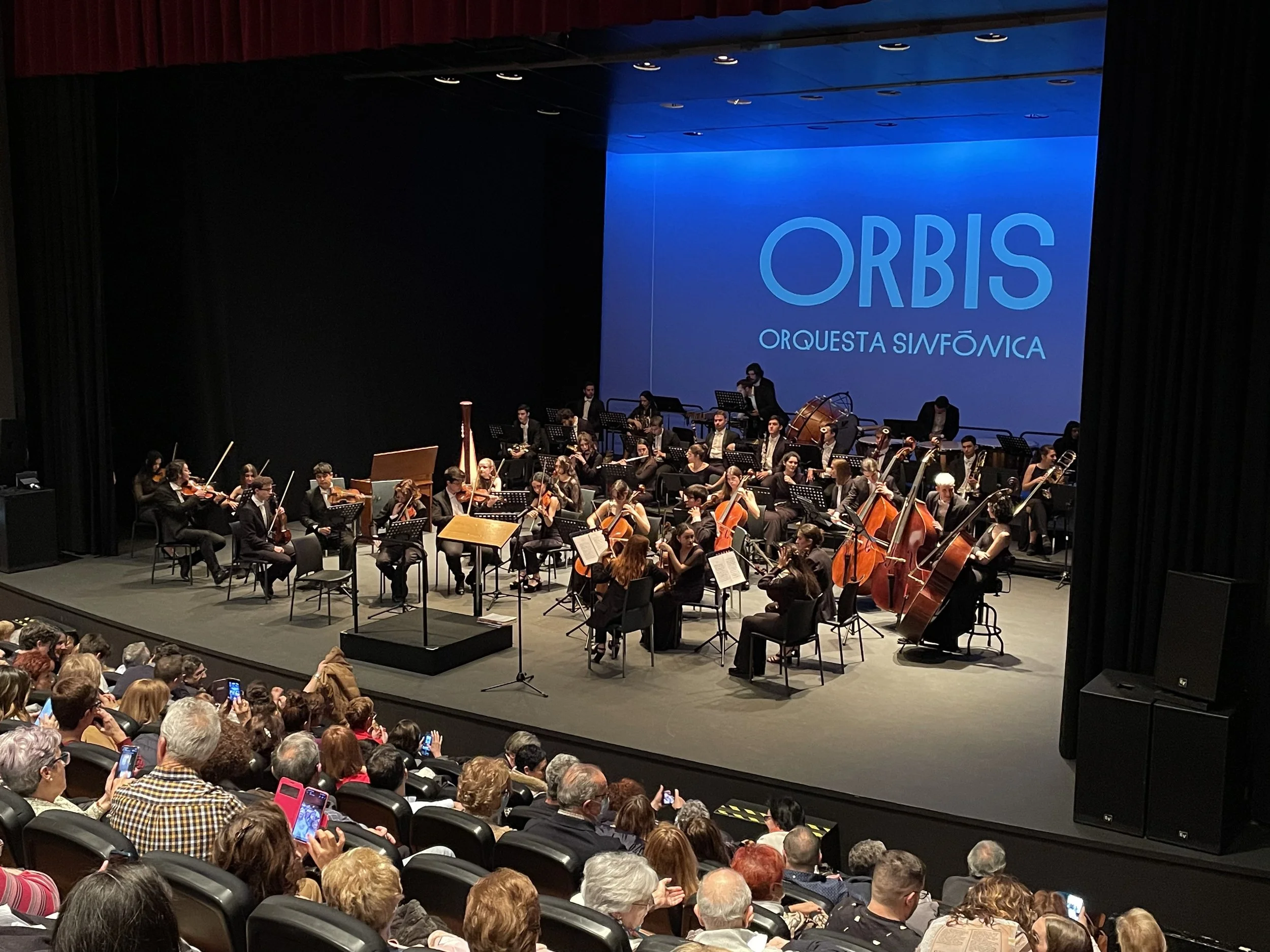 Orquesta sinfónica en un escenario con una pantalla que dice 'ORBIS ORQUESTA SINFÓNICA', con músicos tocando violines, cellos y otros instrumentos en un concierto, frente a un público sentado en la sala de conciertos.