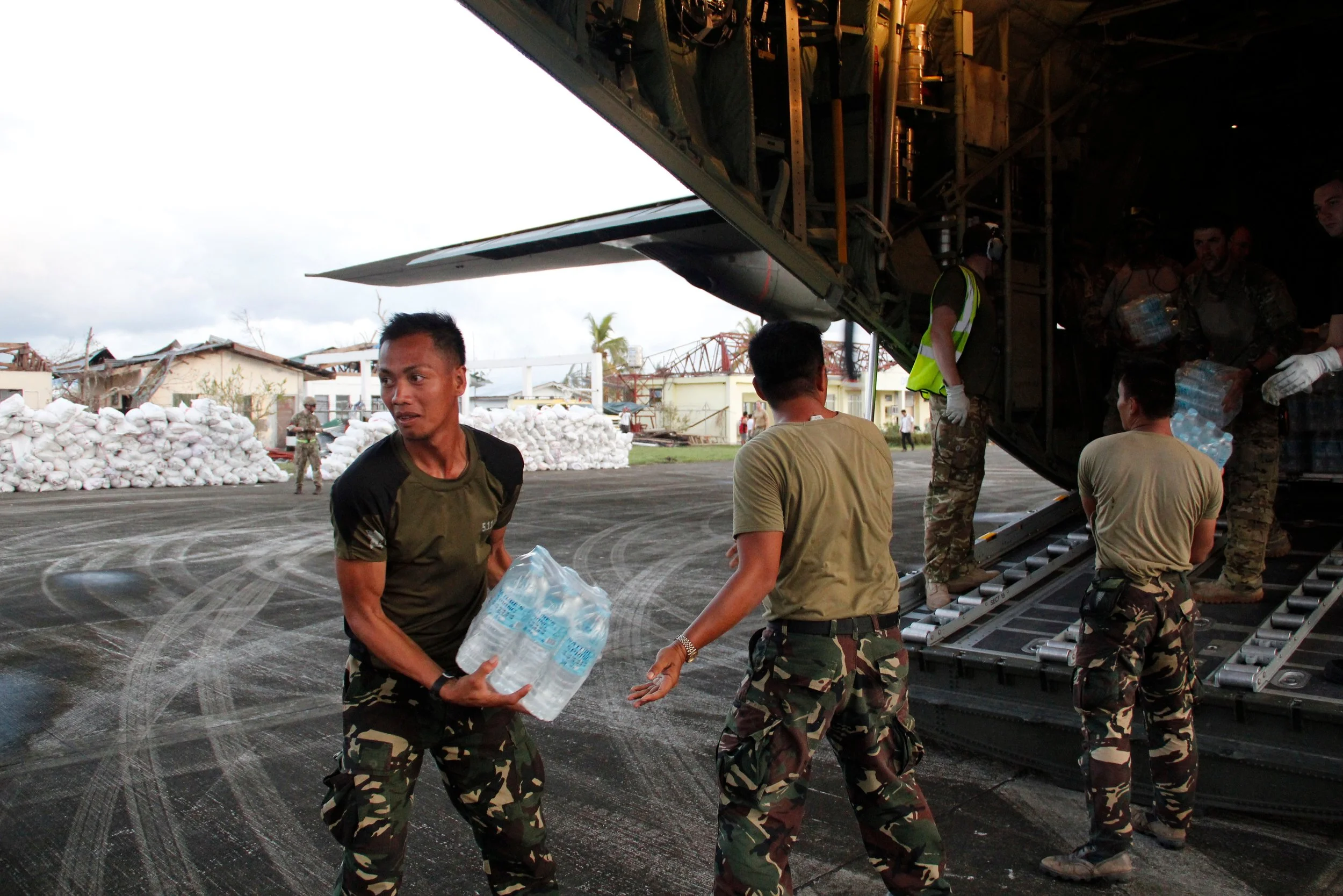 Military personnel unloading bottled water from a plane, with some individuals in the background and damaged structures visible.