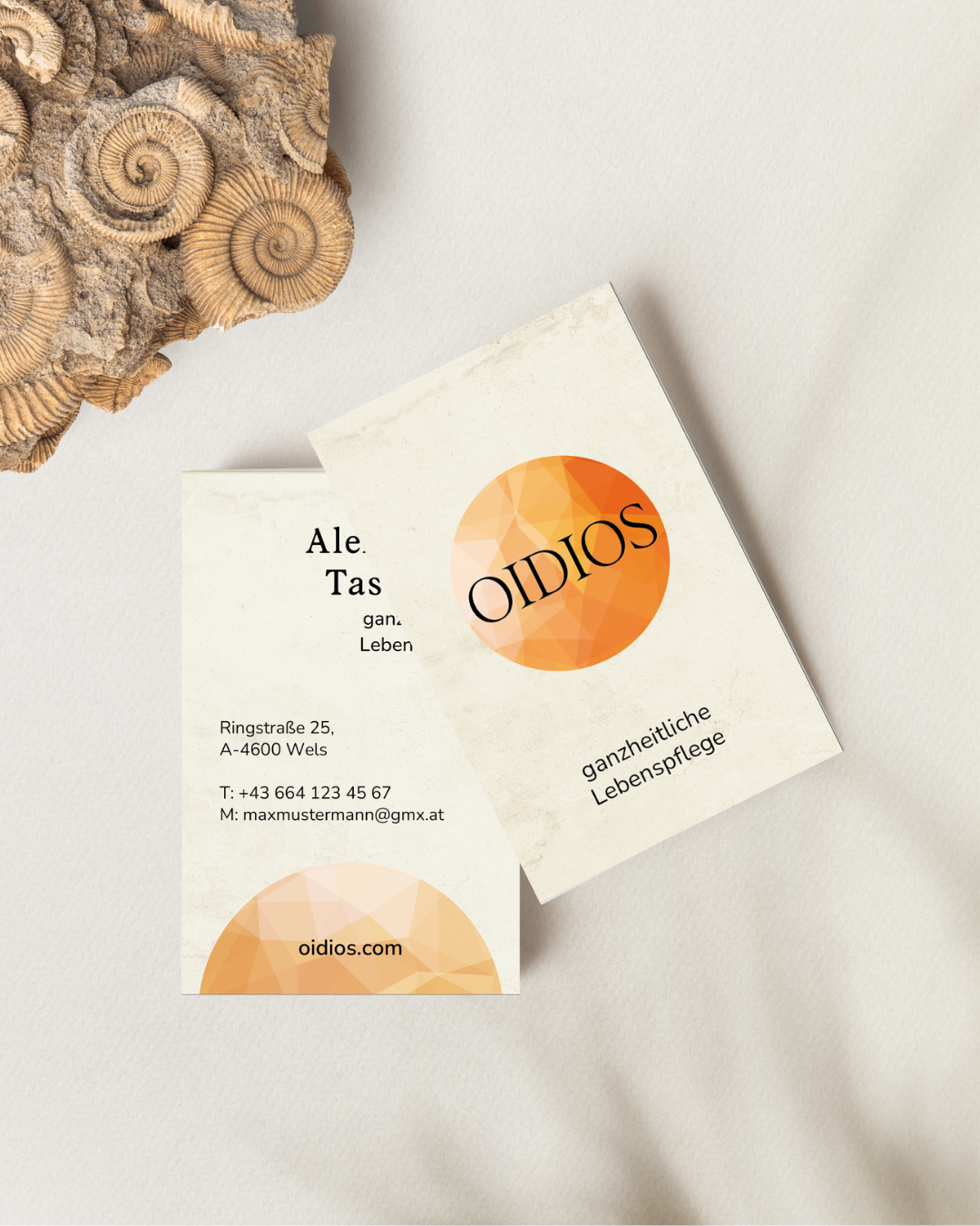 Zwei Visitenkarten mit dem Logo "OIDIOS" und der Aufschrift "ganzheitliche Lebenspflege" auf einer neutralen Oberfläche. Auf einer der Karten stehen Name, Adresse in Wels, Telefonnummer und E-Mail. Links oben sind Ammonitenfossilien zu sehen.