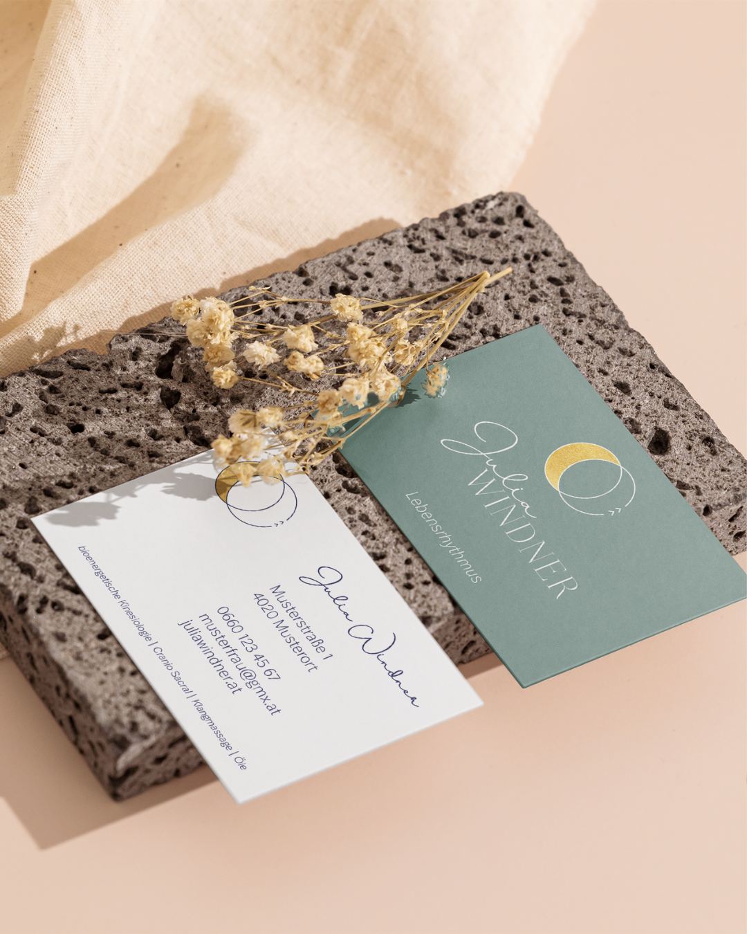 Zwei Visitenkarten liegen auf einem porösen Stein. Eine Karte ist hell und die andere in einem grünen Farbton. Auf der hellen Karte steht ein Name und Kontaktinformationen. Ein Bündel getrockneter gelber Blumen liegt dekorativ auf den Karten.