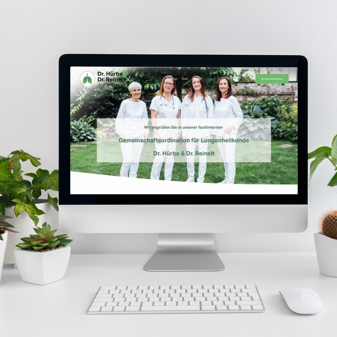 Ein Computerbildschirm zeigt eine Website von Dr. Hürbe & Dr. Reinelt, einer Gemeinschaftspraxis für Lungenheilkunde, mit einem Bild von vier Menschen in weißen Arbeitskleidern im Freien. Auf dem Schreibtisch stehen Pflanzen und eine Tastatur.