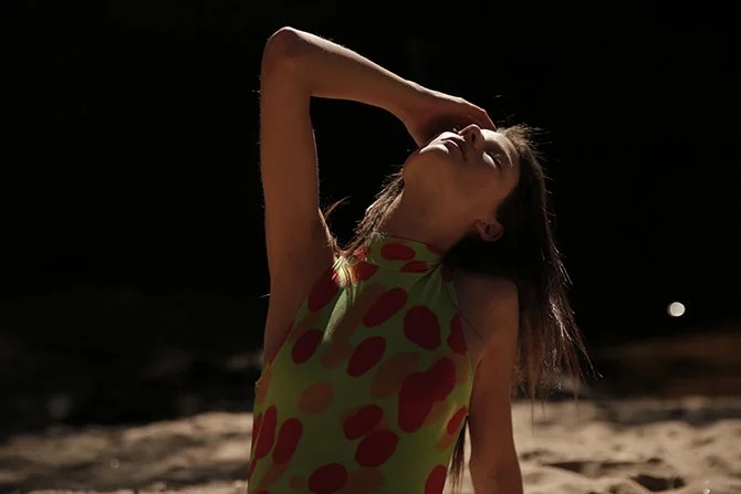 A woman in a colorful dress with red spots on a green background stands on a sandy beach at night, striking a pose with one hand behind her head and eyes closed.