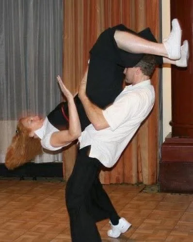 Two people performing a dance lift indoors on a hardwood floor, with curtains in the background.
