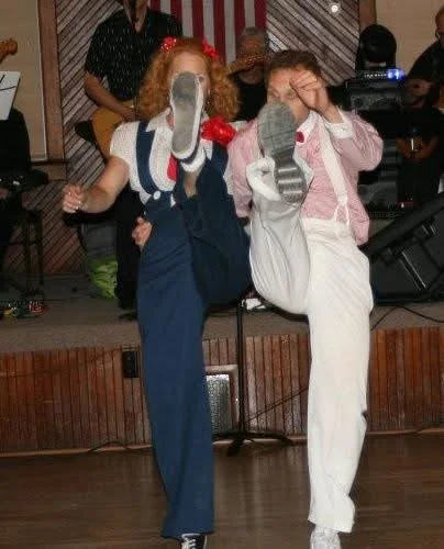 Two people dancing enthusiastically with high kicks on a stage; one wears a blue outfit, the other a white and pink outfit. A live band plays in the background.
