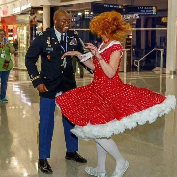 A man in a military uniform smiles while dancing with a woman in a red polka dot dress, with the dress twirling out. They are in a brightly lit indoor space with shiny floors and people in the background.