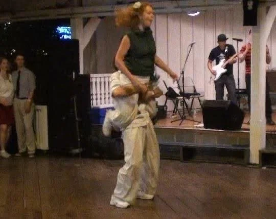 Two people energetically swing dancing on a wooden dance floor, with a band playing live music on stage in the background.