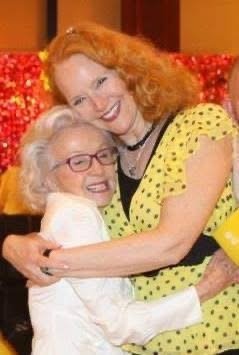 Two women smiling and hugging at an indoor event, with one wearing a yellow dress with black polka dots and the other wearing a white jacket.