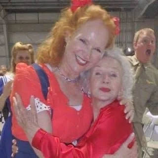 Two women in vibrant outfits, one with red hair and wearing a red dress, the other with white hair and also in red, smiling and embracing each other in a joyful setting. There are people and industrial elements in the background.