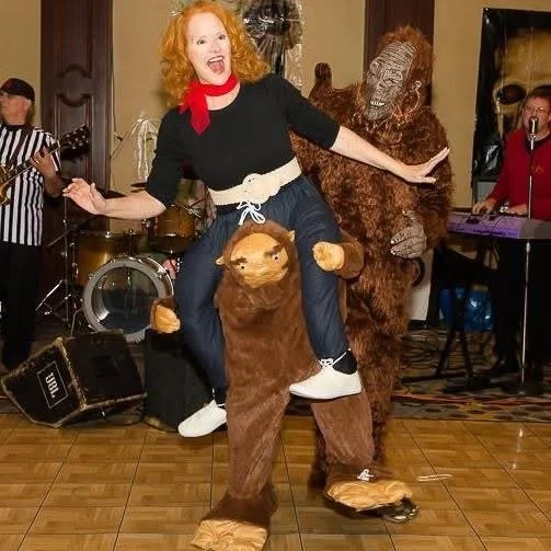 A person in a red wig and black outfit, wearing a humorous costume that appears as if they are riding on an animal, with another person dressed in a gorilla or bigfoot costume. They are in a room with musical instruments and performers in the backgro